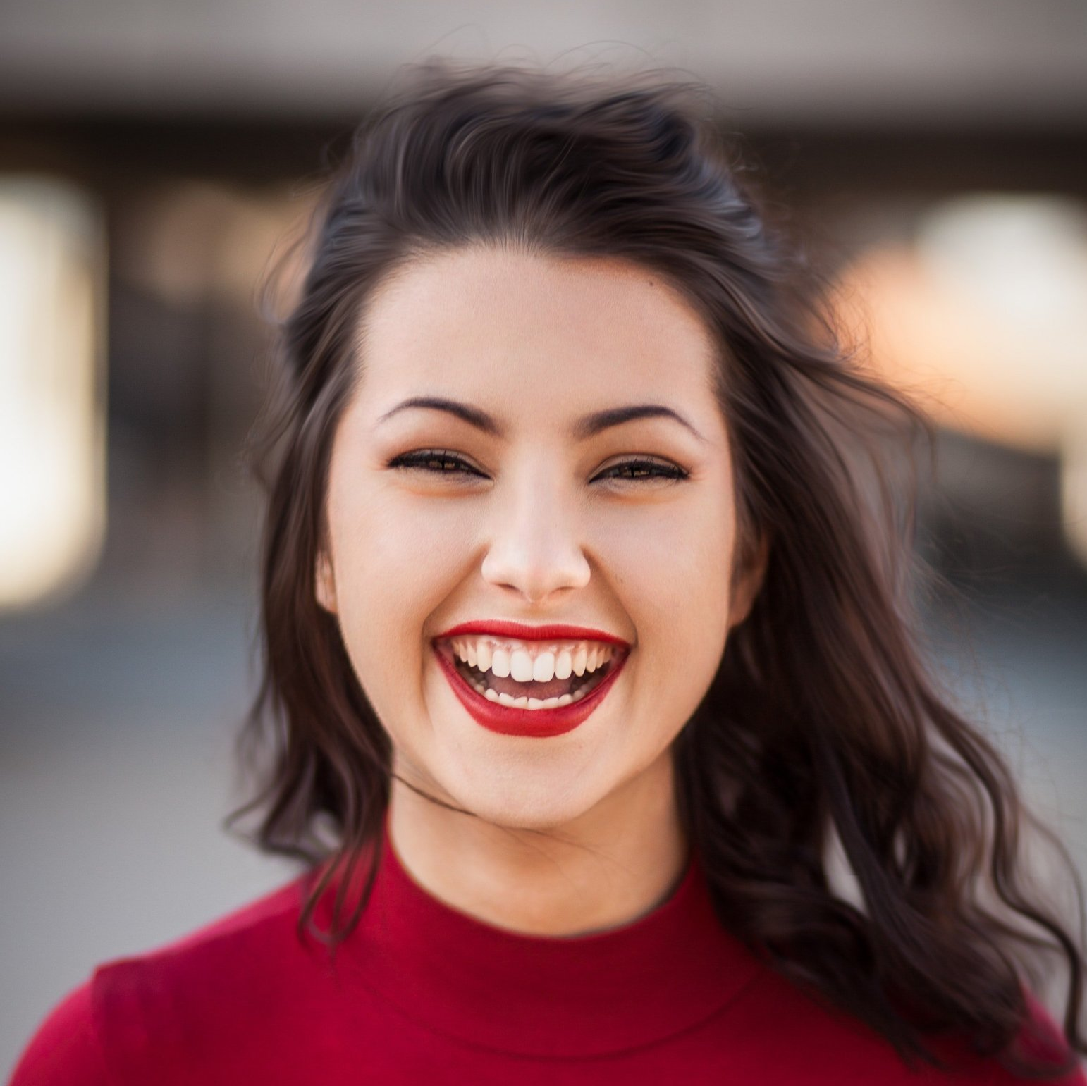Woman with dark hair smiles widely, wearing a red top and lipstick. Outdoors, blurred background.