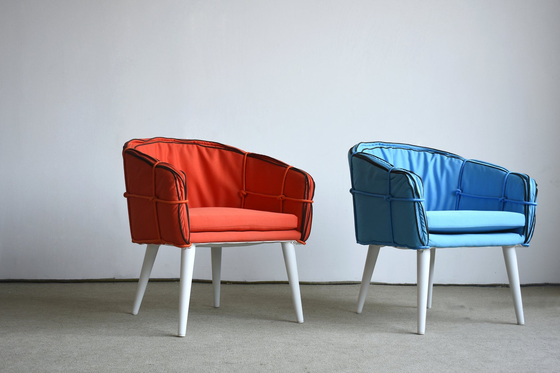 Two modern armchairs, one orange and one blue, with white legs and a stitched design, against a white wall.