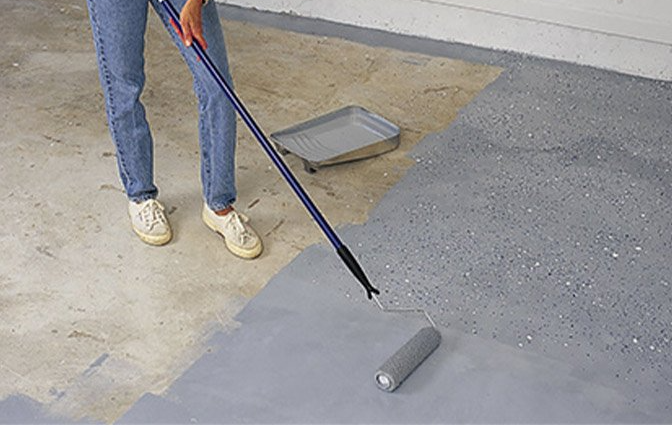 Person painting a concrete floor gray with a roller and tray.