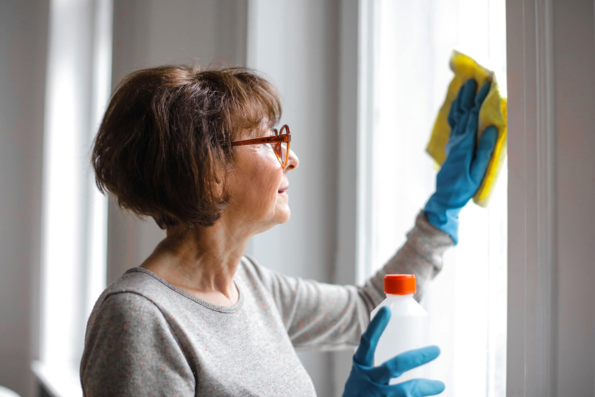 Woman wearing glasses and gloves cleaning a window with a yellow cloth and cleaning solution.